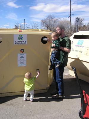 Sarah reaches up with her can. David gives Noah a boost to recycle.